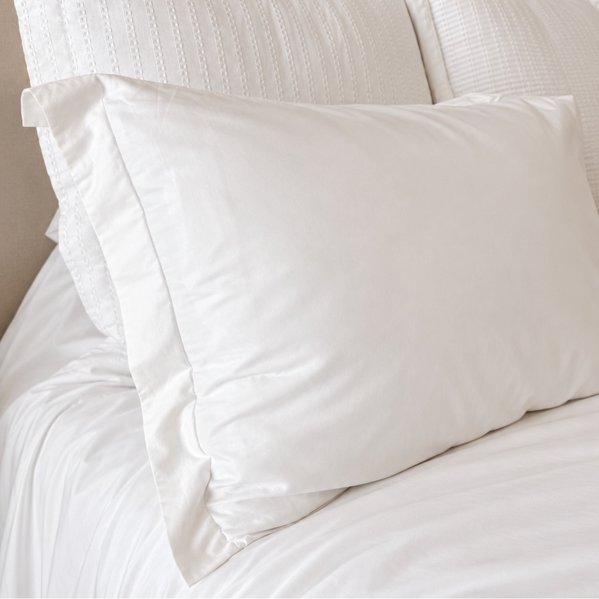 A close-up of a white Heritage Collection pillow shame resting on white bedding, with textured white pillows and a beige headboard in the background.